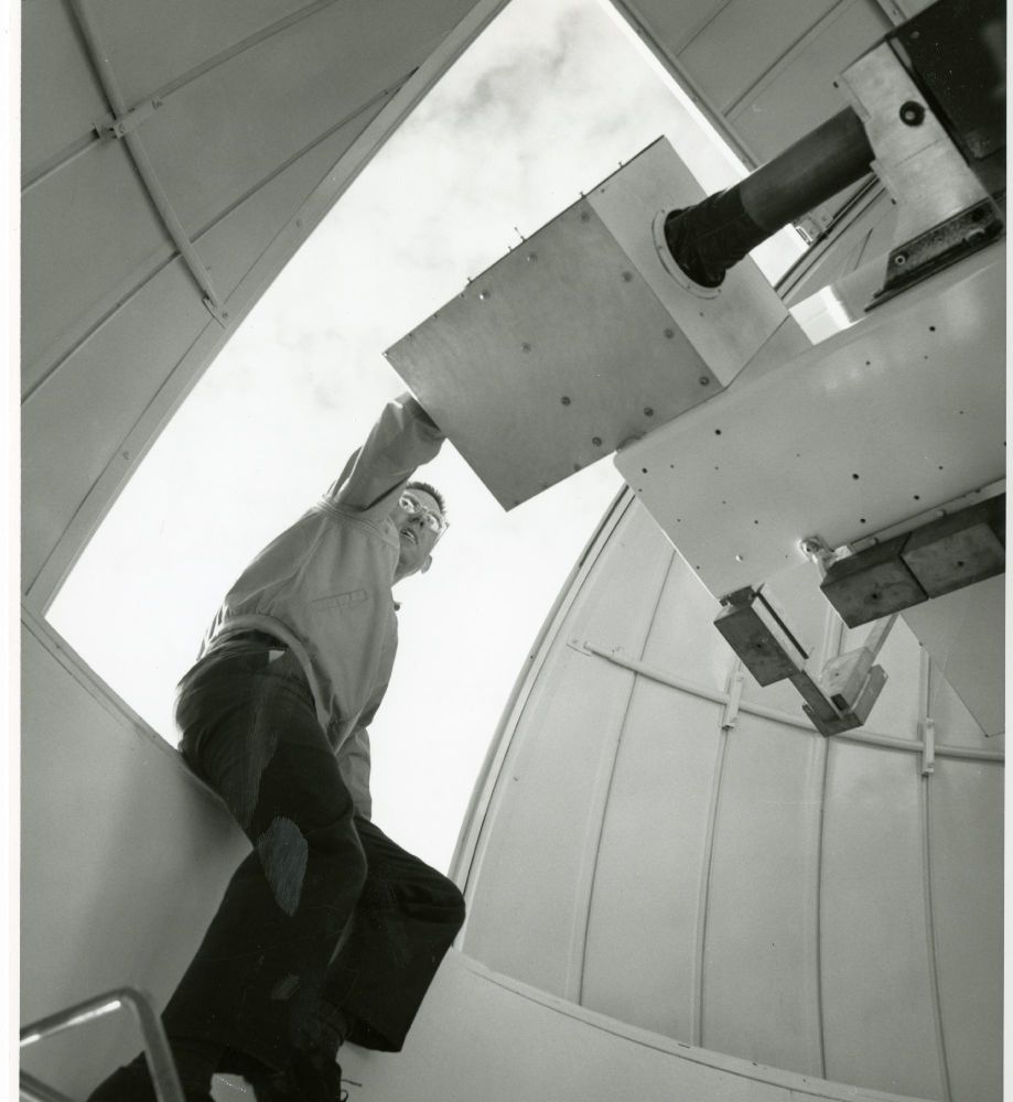A person is adjusting a large telescope inside an observatory dome, reaching upward towards the telescope's mechanism. The background shows a clear sky through the dome's open panels.