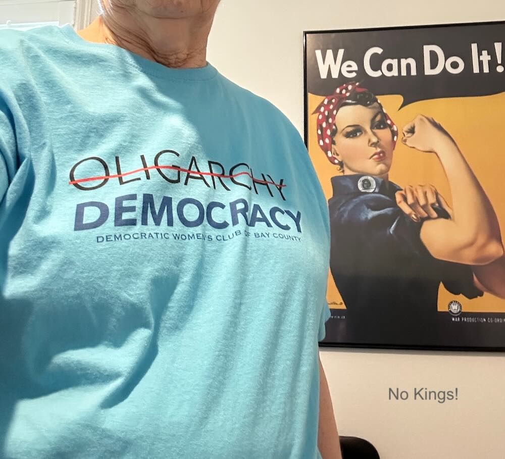 An old woman in a shirt with the word Oligarchy crossed out and Democracy written under it. Behind the woman is a Rosie the Riveter sign saying We Can Do It!