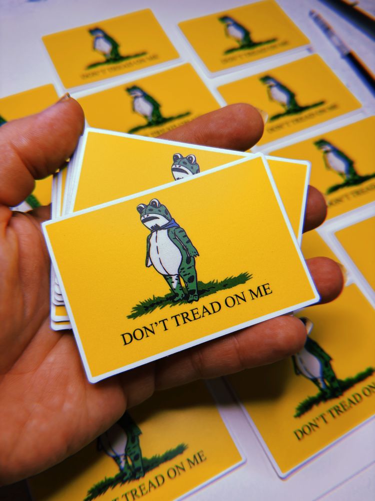 A hand holding a yellow sticker with the Portland frog and the text DONT TREAD ON ME 