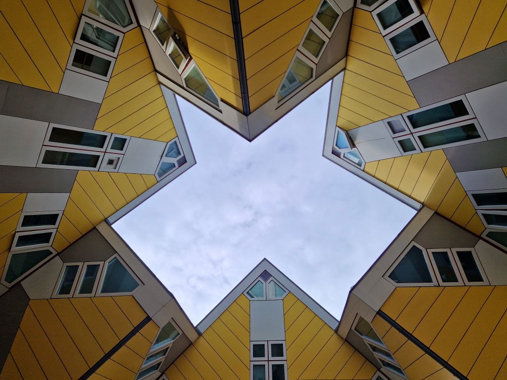 Gelbe, würfelförmige Häuser von unten fotografiert, sodass sie das Bild umrahmen. In der Mitte eine Öffnung zum bewölkten Himmel.