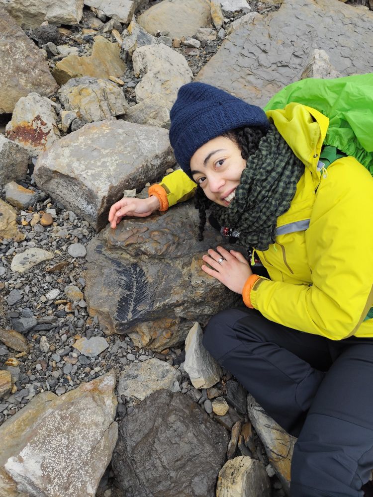 Sara wearing brightly coloured hiking gear crouched next to a large fossil plant (the size of a forearm).