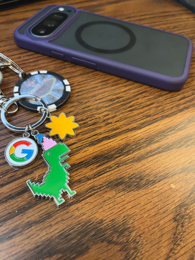 My Pixel 9 Pro (sage) in a transparent case with purple edges lying face down on a table of dark wood, next to the birthday keychain from Google that presumably caused the shipping delay