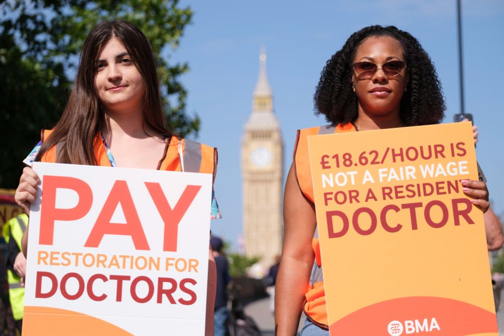 Two doctors in orange hold protest signs in front of Big Ben. One sign reads "Pay restoration for doctors" and the other says "£18.62/hour is not a fair wage for a resident doctor"
