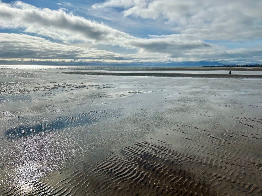 An empty beach at low tide with rippled wet sand. There are mountain peaks on islands in the distance, the sky is blue and there are big white pale grey clouds.
