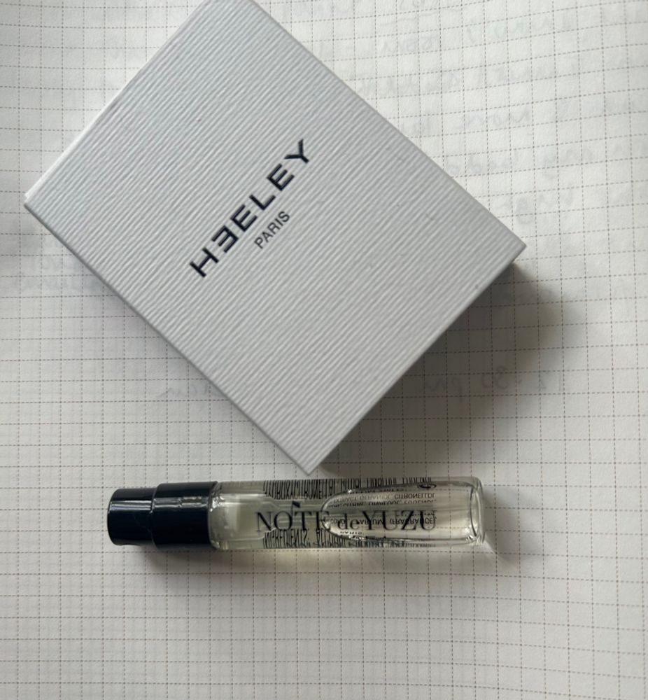 A glass sample bottle of Note de Yuzu by Heeley Parfums is lying on grid pattern notepaper. There is a small white slightly ridged box with the Heeley Parfums branding on.























