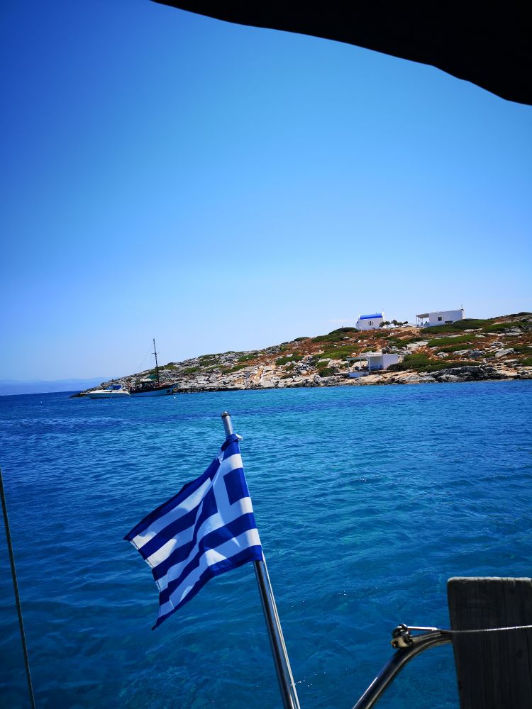 Die griechische Flagge im Wind die an einem Segelboot befestigt ist. Man sieht blaues Meer, blauen Himmel und Hühel mit Gebäuden.