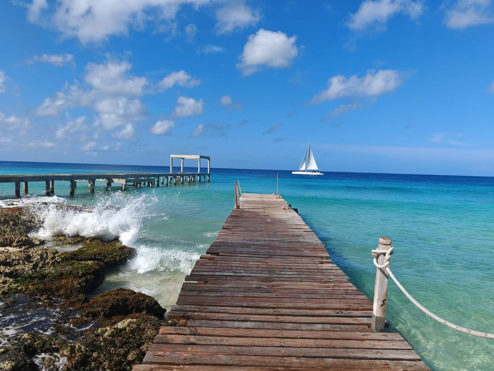 Blauer Himmel mit Wolken, Meer in n verschiedenen Blautönen, ein Segelboot in der Ferne, 2 Stege die ins Meer führen.