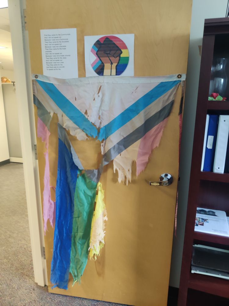Office door with tattered and shredded progress pride flag hanging vertical and progress pride flag with multiracial fist (printed) above.