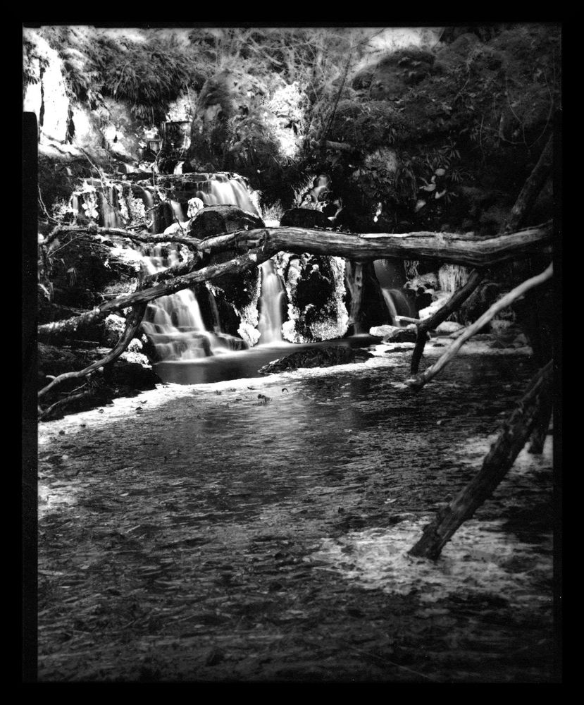 Black & White image taken on a 4x5 camera. 

A stream is partially frozen, the ice is flows down a small cascade, in between the water flows smoothly. The foreground is a frozen pool of water with a fallen tree branch across it.