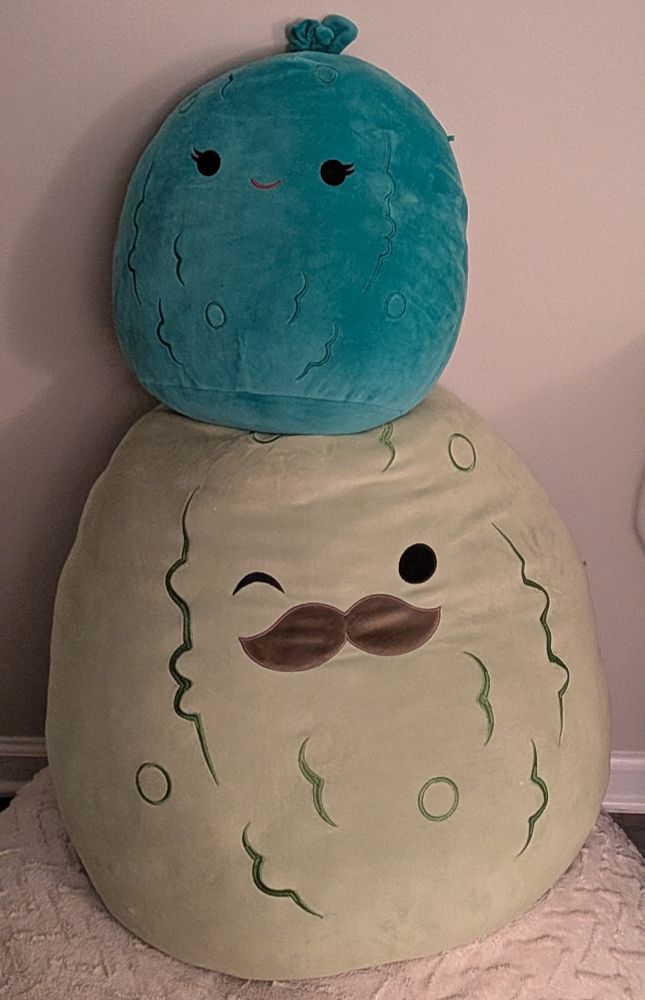 A smaller (14 inch) Squishmallow pickle sitting on top of a larger (24 inch) Squishmallow pickle. The larger pickle has a moustache and is winking.