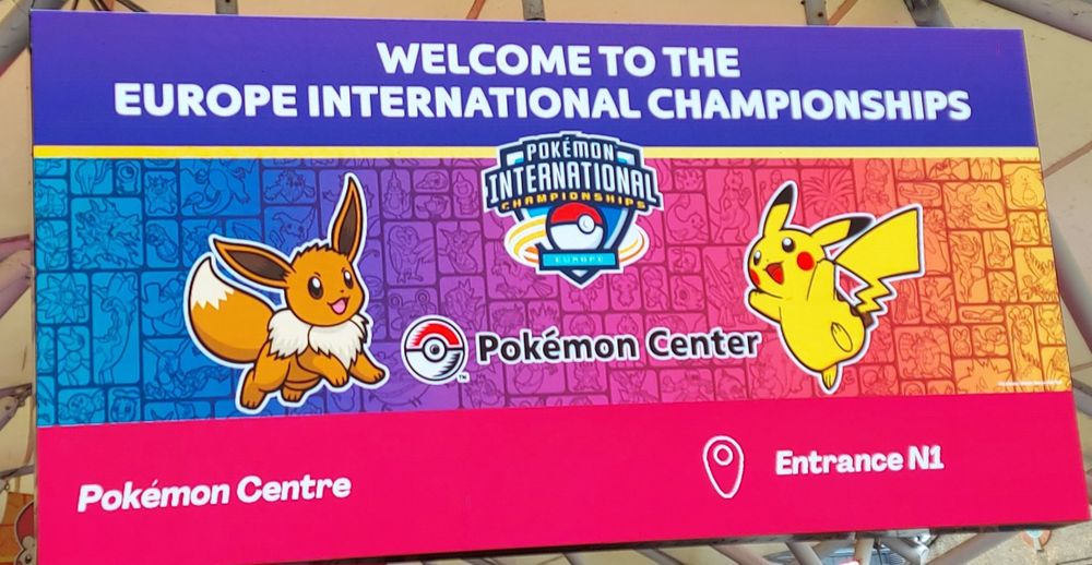 A large screen sign that says 'welcome to the  Europe International championships!' With a Pokemon International logo in the center. Eevee and Pikachu are pictured in front of a rainbow gradient background, as well as text for directions to the pokemon center. 