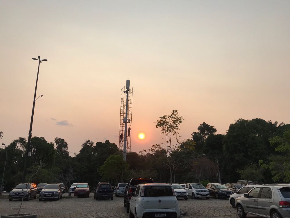 Pôr do sol em um estacionamento da UFAM. Horizonte fechado com árvores. Em uma torre de celular homens trabalham.