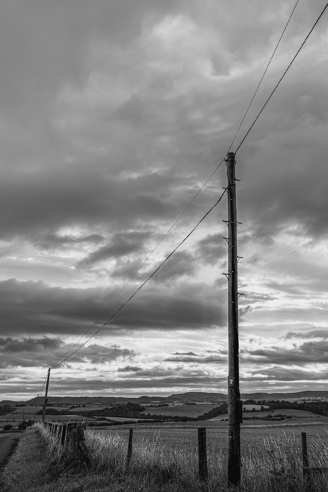 A black and white photograph of a telegraph pole with wires a fence leads down to another pole with a field to the right side. The sky is dark and stormy. 