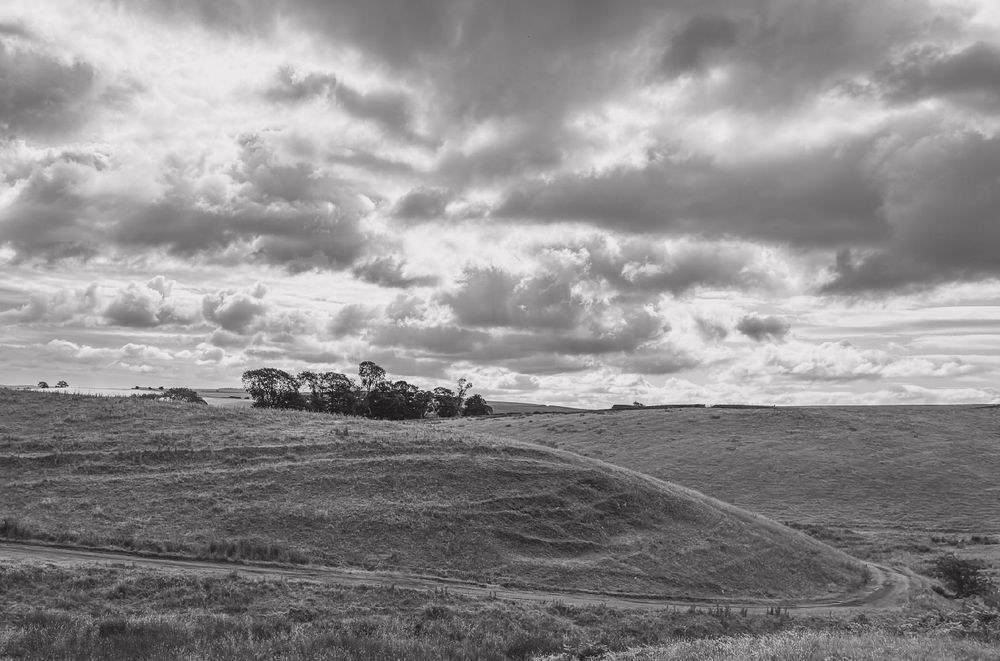  A black-and-white photograph of two small hillocks with a cloudy sky above. Between them is  a small wood and in front of the first hillock is a footpath winding between the two. Across the hillsides are the marks of mediaeval ploughing. 