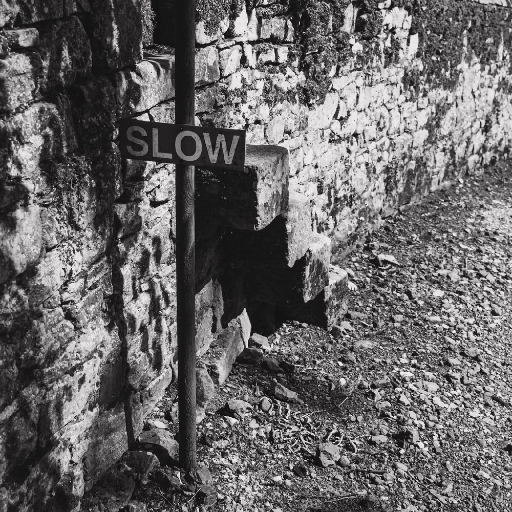 A black and white photograph of a stone wall with stone steps leading over it. In front is a sign with the word SLOW in capital letters.
