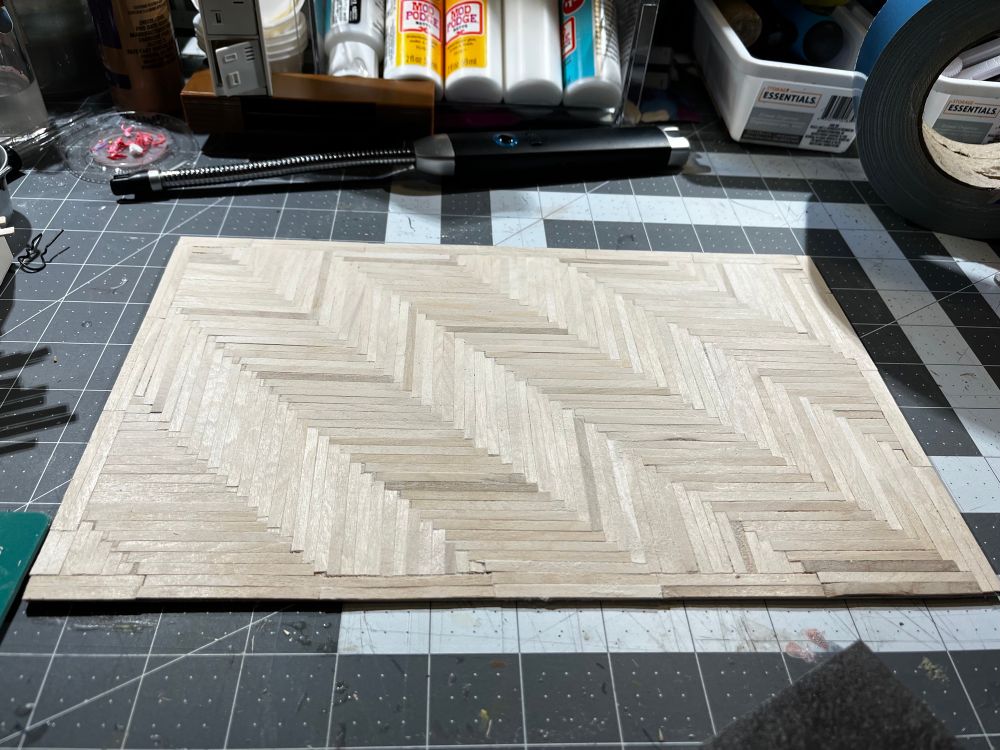 7.5”x11.5” rectangle of unfinished herringbone wood flooring made of individual strips of cut wooden coffee stir sticks. 