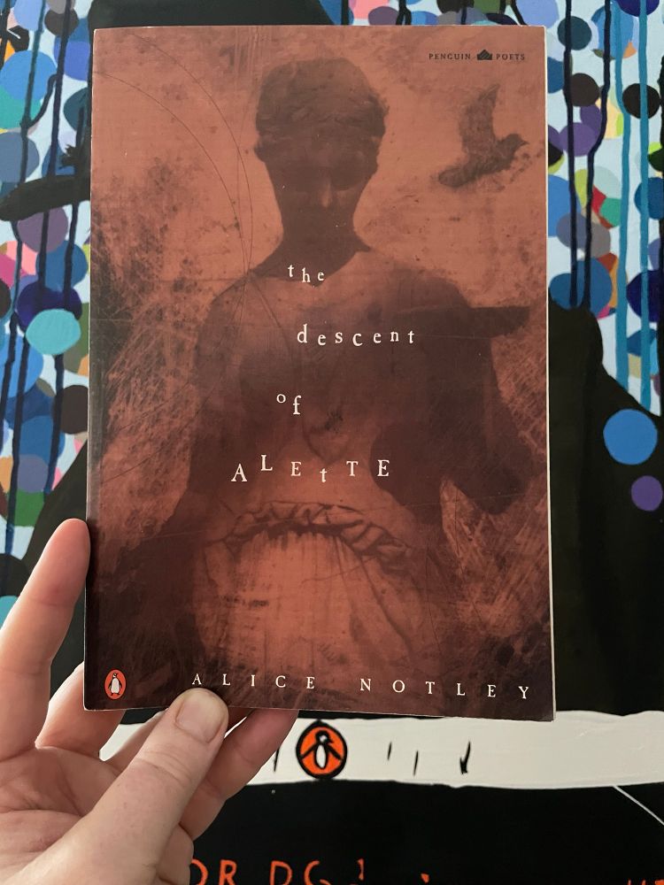 An orange, white, and black book and its cover is shown. The book’s title is The Descent of Alette by Alice Notley. 