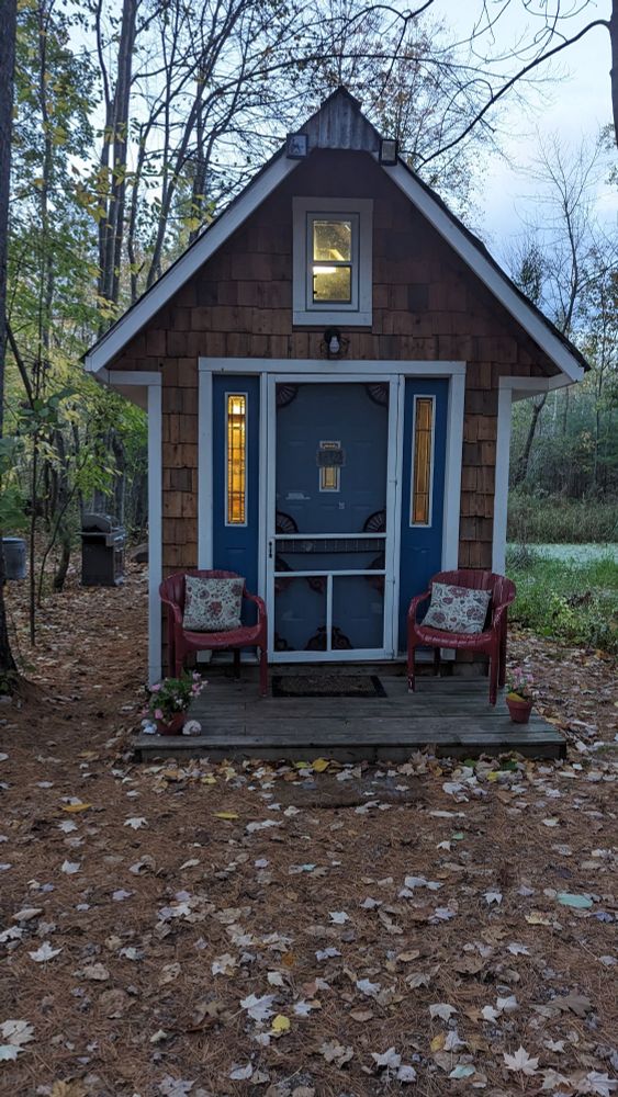 Photo shows a small, wooden tiny house with a blue front door and red chairs on a small deck.