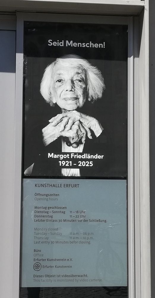 Eingang der Kunsthalle Erfurt.
Rechts der Tür über dem Schild mit den Öffnungszeiten ein SW-Porträt einer alten Dame mit weißen Haaren, die direkt in die Kamera blickt und vor ihrer Bust ihre Hände zusammen führt.
Oben steht weiß auf Schwarz: "Seid Menschen!", unterhalb ihrer Hände: "Margot Friedländer 1921 - 2025".