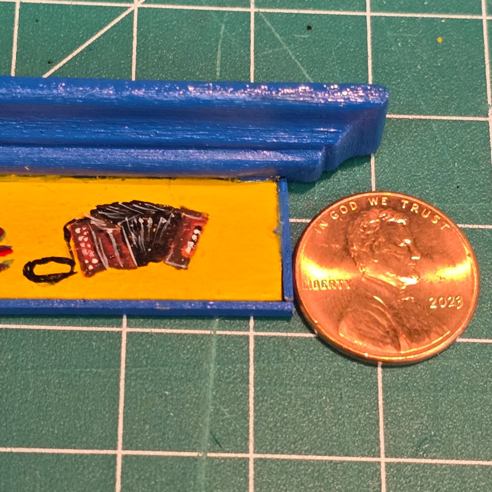 Tiny acrylic painting of a button accordion on a yellow background surrounded by blue moulding. A penny sits next to it for size comparison.