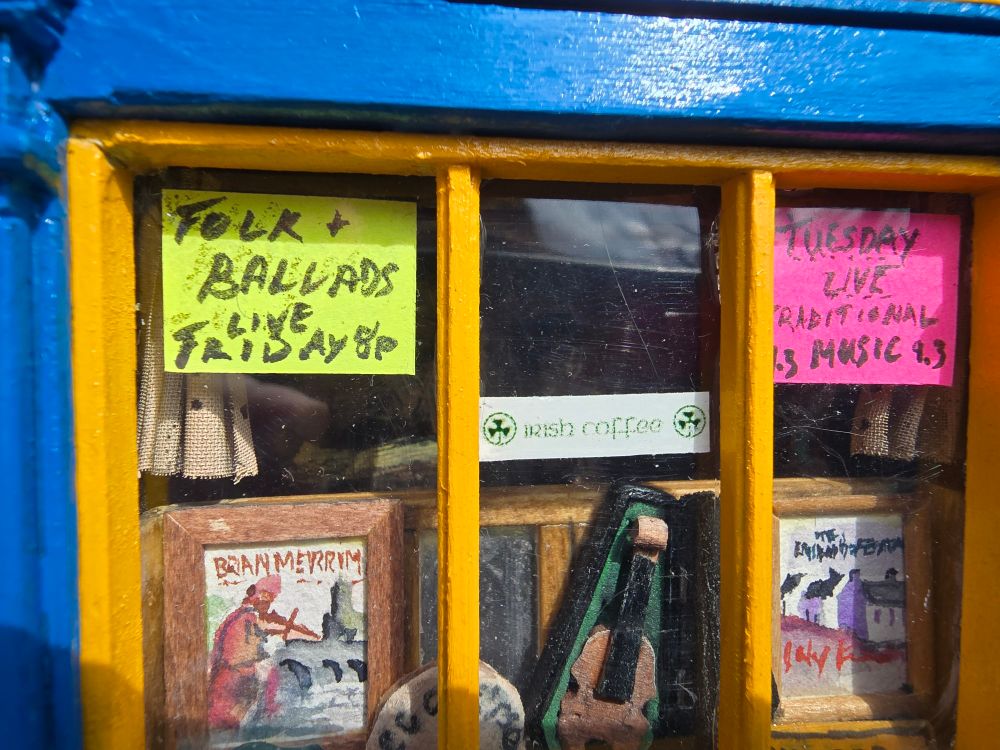 Closeup of the window of the pub featuring handpainted watercolor posters, a scratch built fiddle in its case and other signs and decor.