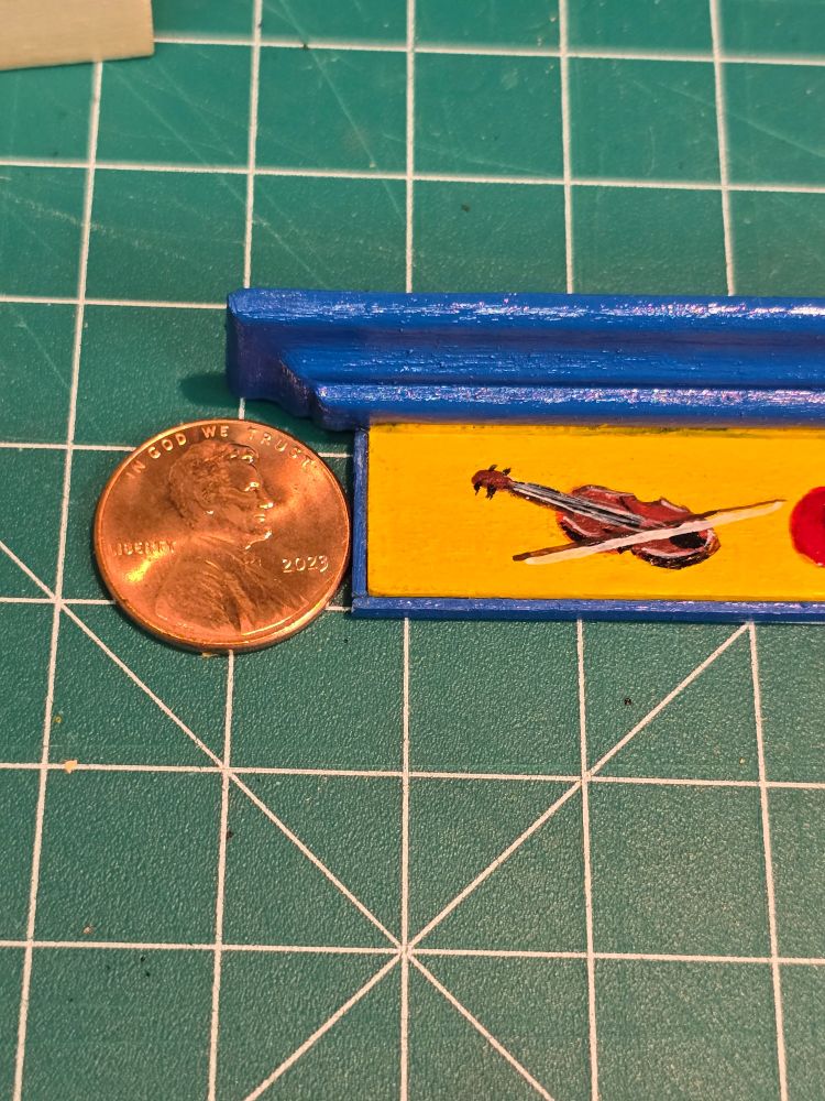 Tiny acrylic painting of a fiddle and bow on a yellow background surrounded by blue molding. A penny sits next to it for comparison.  The fiddle looks to be about the right size for Lincoln to play!