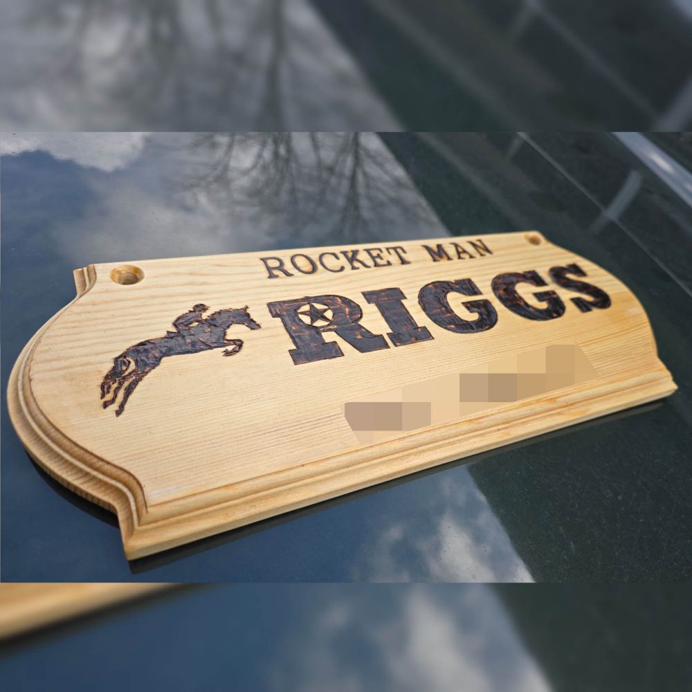 Wooden stall sign which reads the name "Rocket Man" across the top. In the middle, his barn name "Riggs" is displayed in a bold, serif font with a star in the eye of the letter "R". A silhouette of a jumping horse and rider is to the left of "Riggs". The owner's name at the bottom of the sign had been obscured for privacy.