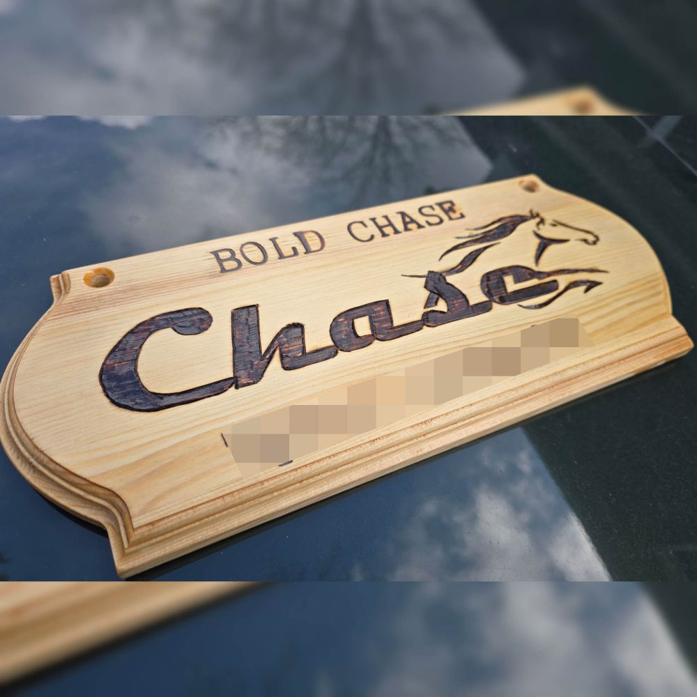 Wooden stall sign which reads the horse's show name "Bold Chase" across the top. In the middle, his barn name "Chase" is displayed in a sleek, Bold, cursive font that gives way to a stylized illustration of a horse at full gallup. The owner's name at the bottom of the sign had been obscured for privacy.