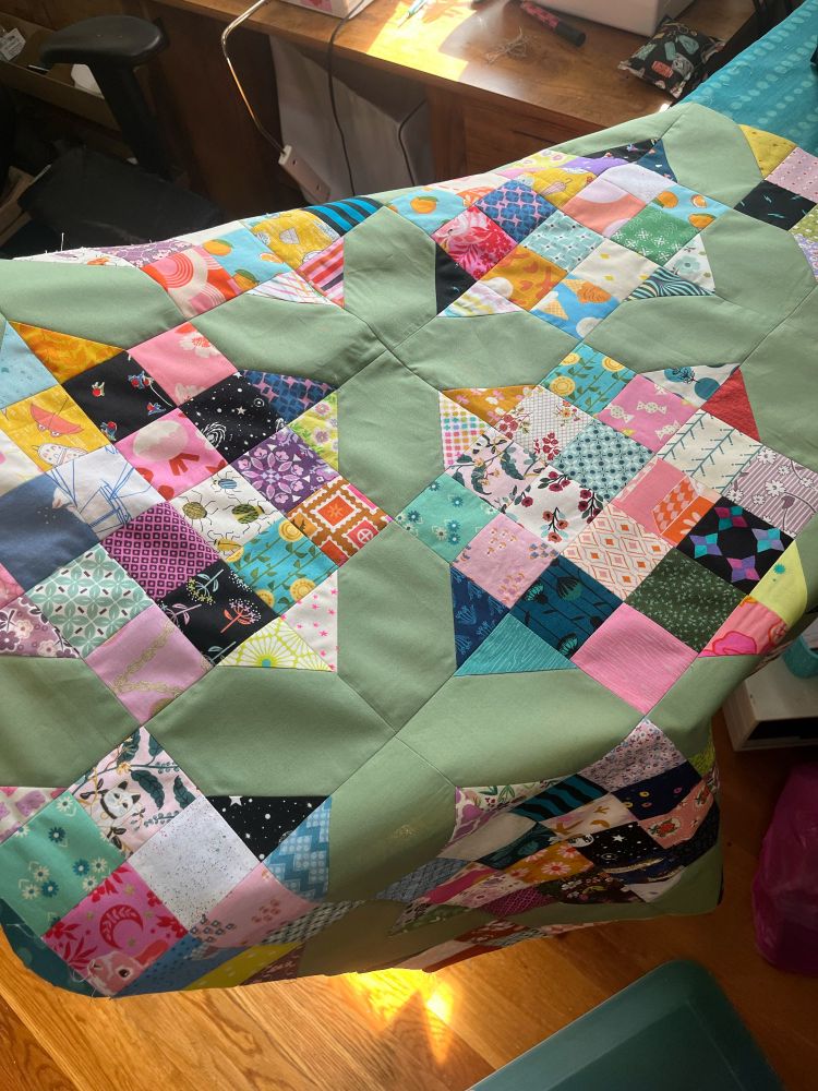 A patchwork quilt in progress- multicolored 2in squares on a sage green background draped over an ironing board. Background is bits of my sewing room
