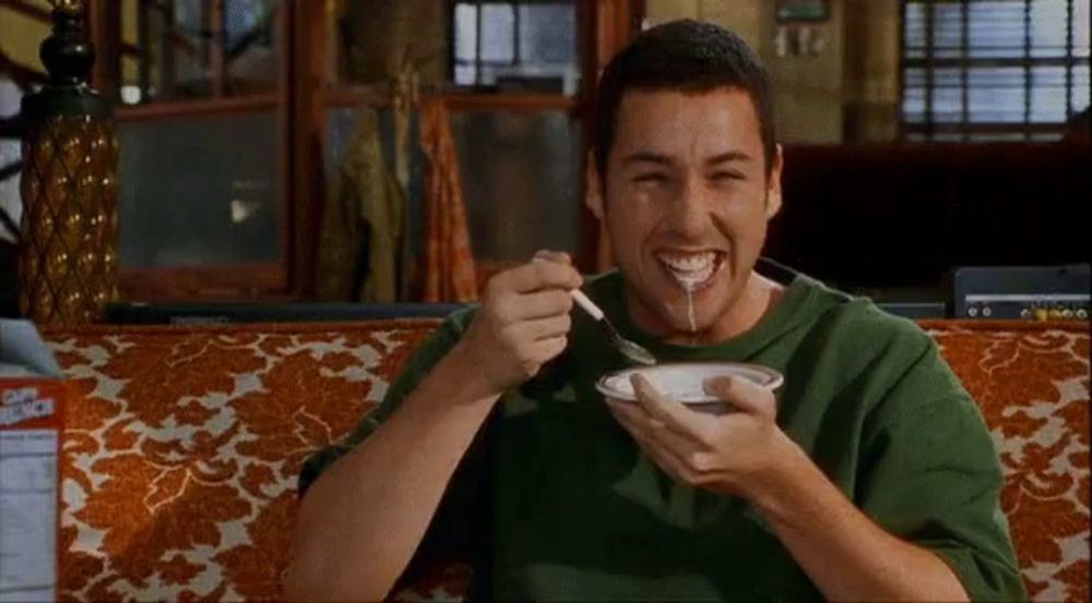 a man is sitting on a couch eating a bowl of food with a spoon in his mouth .