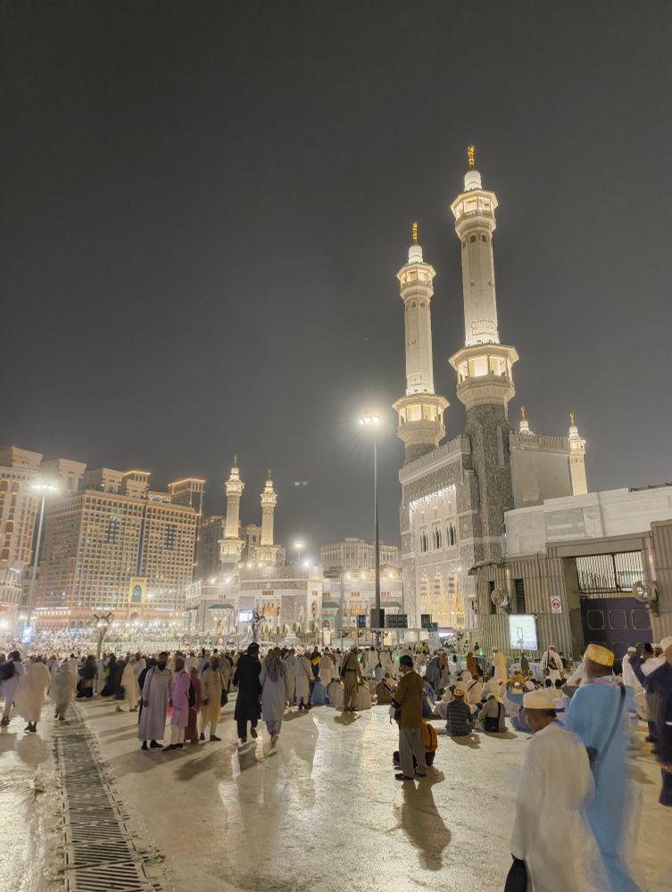 The photo shows Masjid-Al-Haram in Makkah, Saudi Arabia, with lights illuminating the huge mosque. Many people are walking around at night, under a dark sky, and the beautiful white minarets are glowing.