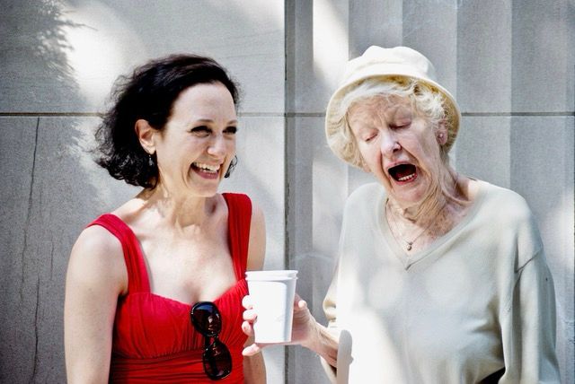 Two white women laughing in the sunshine in front of a light stone building. The younger one with dark hair laughs adoringly at the older, light haired one. The older one is wearing a white hat.