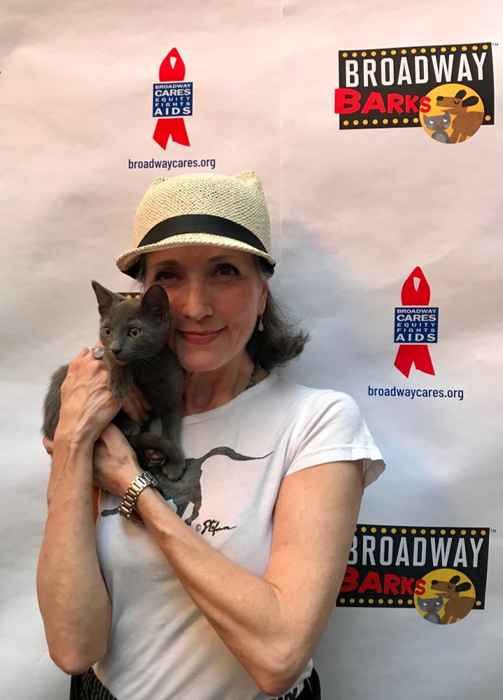 Beautiful little gray kitten held by a dark-haired white woman in a white tshirt and light straw hat. The woman is smiling because she gets to hold a sweet little kitty. The kitten may or may not be smiling. He’s definitely calm. Behind them is a sign saying “Broadway Barks” and Broadway Cares/Equity Fights AIDS.