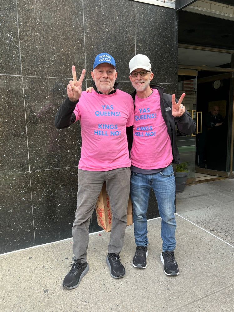 Two smiling men making Peace signs with their hands, wearing pink tshirts that read: Yas queens  Kings hell no. One man is wearing a hat that reads: Mama Gaga. 