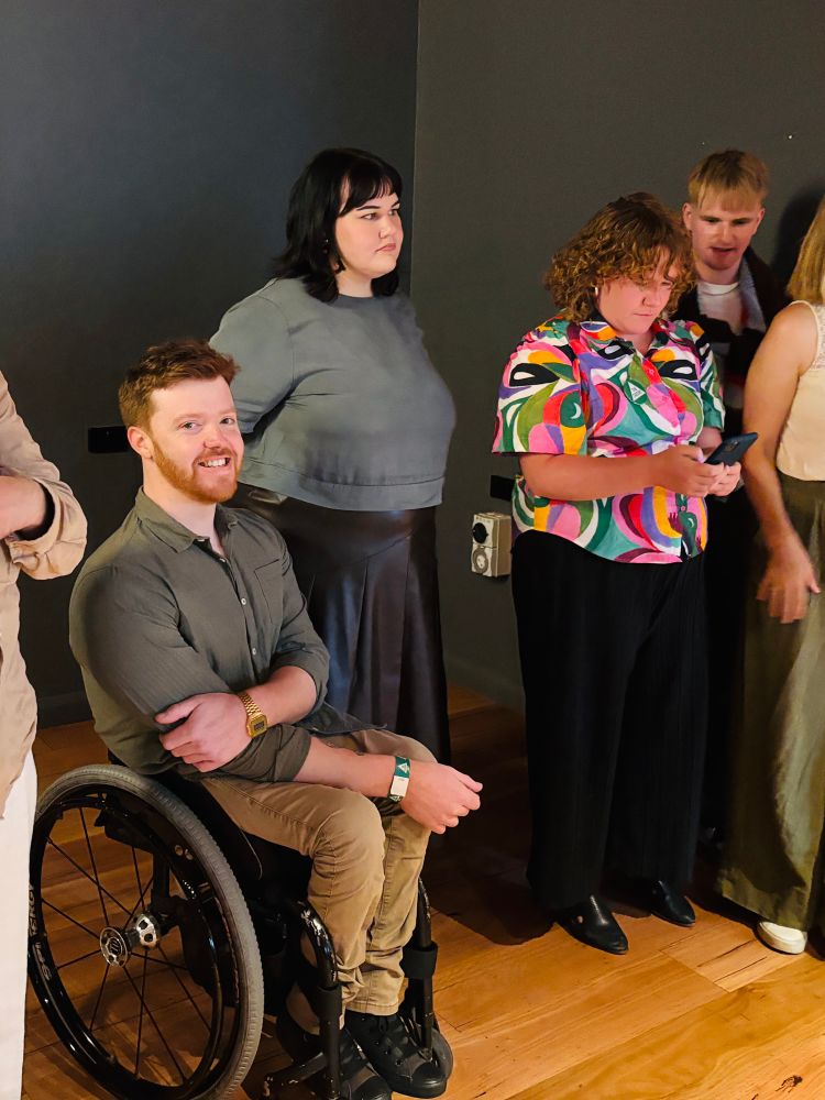 Sam Carter (in wheelchair) smiling and looking at camera. Standing next to him are Dani, who is watching something out of frame to the left, and Isabel who is looking at her phone.