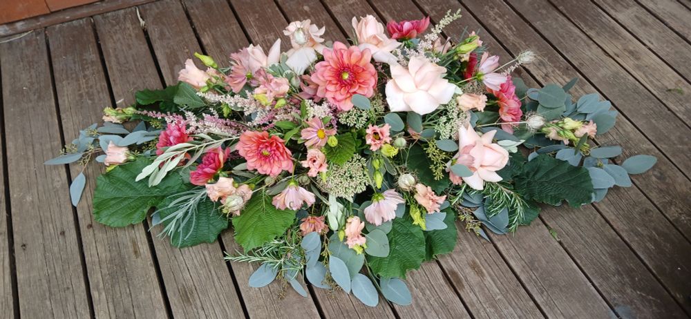 An oblong arrangement of flowers to go on a funeral casket. It includes blush dahlias, sedum, astilbe, roses and lisianthus in various shades of blush pink and pale yellow, with greying green eucalyptus and green beech leaves.