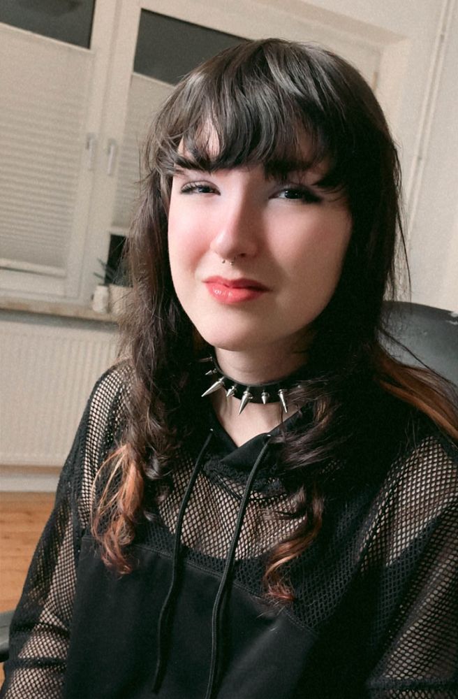 Cute Girl with a spikey chocker sits in her gaming chair with a mesh top. She is smling and has a septum