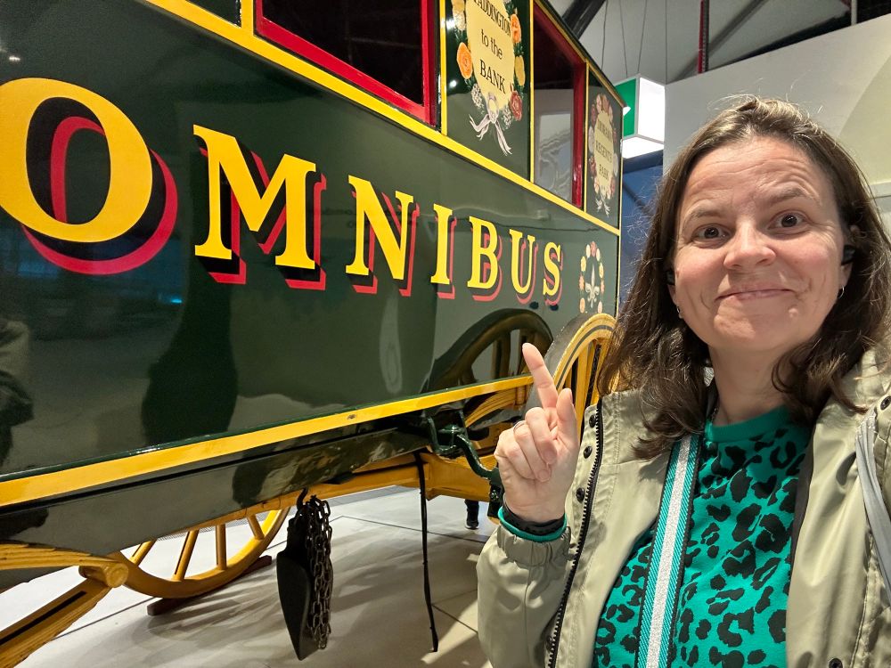 A selfie of a person standing in front of a large wooden omnibus. It is painted dark green, with the words ‘omnibus’ painted in large yellow letters along the side.