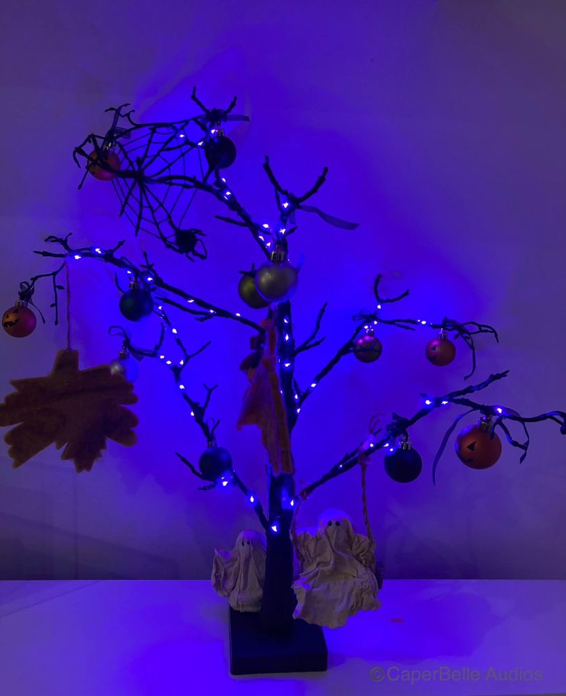Halloween spooky tree decoration with purple fairy lights that give an eerie blue glow, autumn leaves, spider and web, pumpkin baubles, a ghost playing on a swing and another ghost hiding behind the trunk. 
