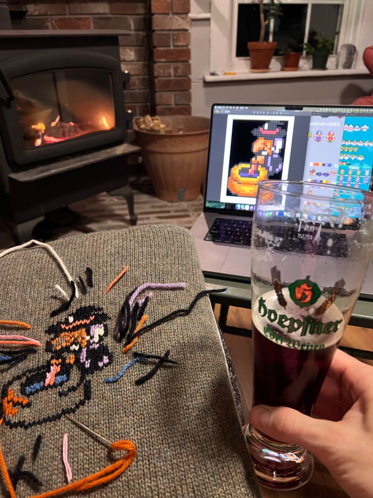 Scene of cushion being embroidered with witch from Zelda Link to the Past, beside hand holding a glass of beer and laptop showing pattern and wood stove burning in background