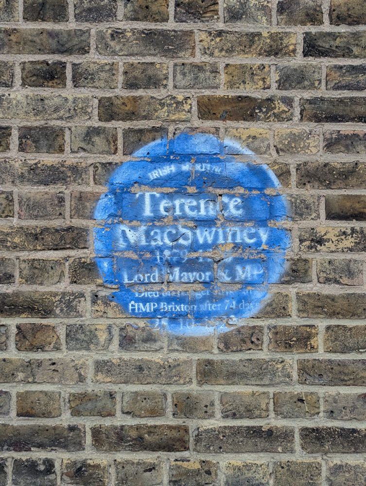 Faded painted white text on faded painted blue background on brick, in the form of a standard blue plaque - some of the text is illegible but the subject of the plaque is Terence MacSwiney.