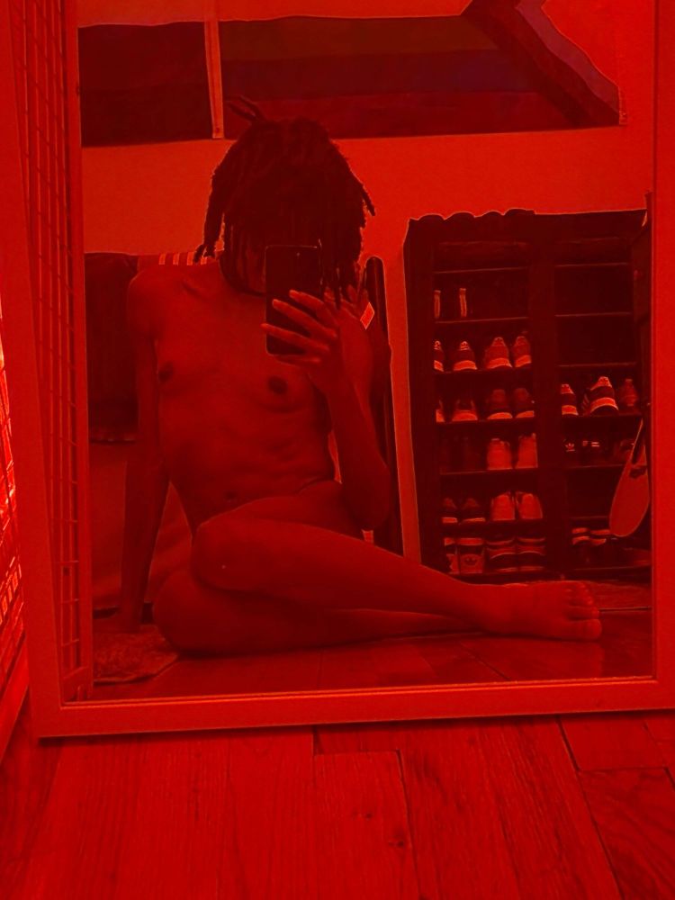 Picture of Hyena sitting on the floor, covered in a red hue. Her body is fully nude as she shits in front of a mirror, her legs crossed in front of her, hiding her face with her hair and phone. 