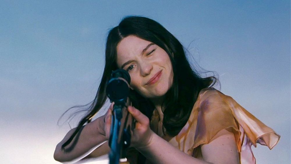Mia Wasokowski, in a shot from the film Stoker. She is a young white woman with dark brown hair, wearing a gauzy dress, squinting down the barrel of a rifle