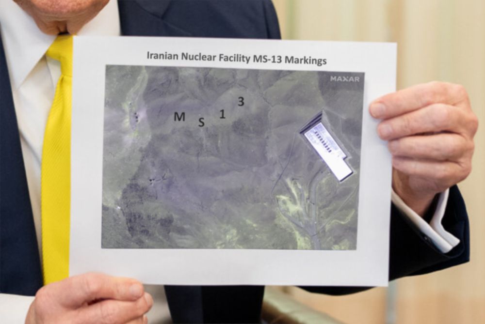 Donald Trump holding up a piece of paper with a poor quality printout: the text above reads "Iranian Nuclear Facility MS-13 Markings", the image below is a satellite photo of one of Iran's bombed nuclear sites, with "M S 1 3" crudely written in black Calibri font over the top