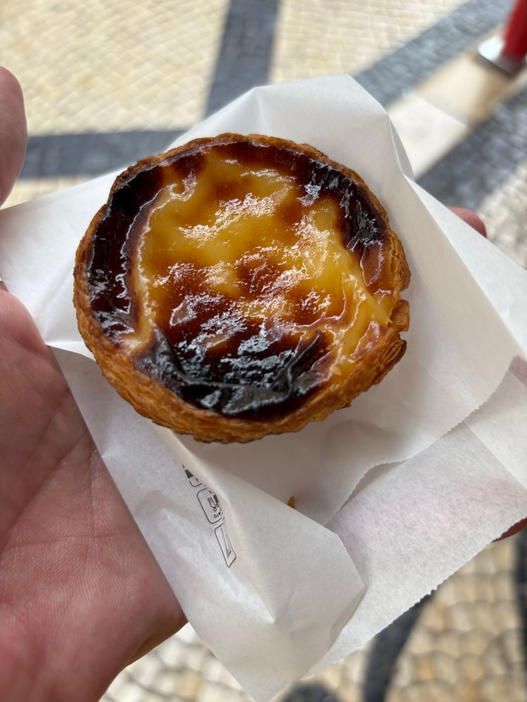 A hand holding a flattened small paper bag on which sits a pastel de nata