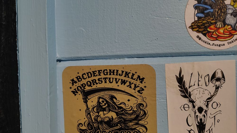 top 2 3rds of a band sticker with a grim reaper girl in the middle and a ouija-soard style alphabet above it. would kind of rule as a band name tbh lol