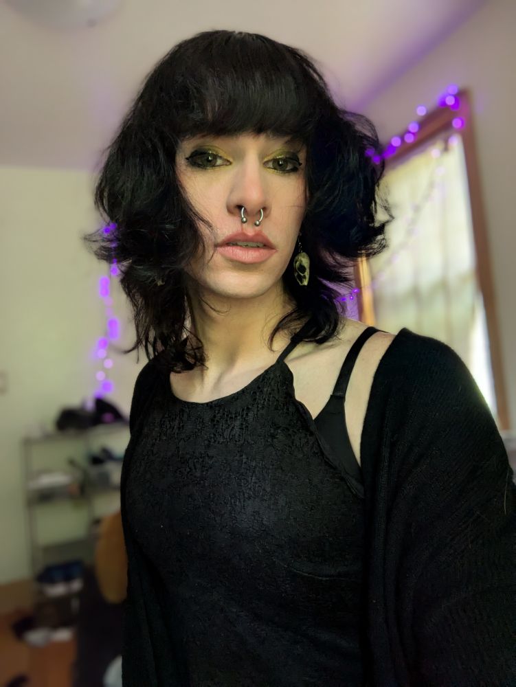 Hex, in a black dress and cardigan, sitting on her bed.