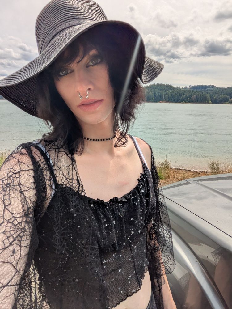 Hex in a spiderweb shawl, constellation crop, and floppy, black sunhat, standing by her car in front of a lake