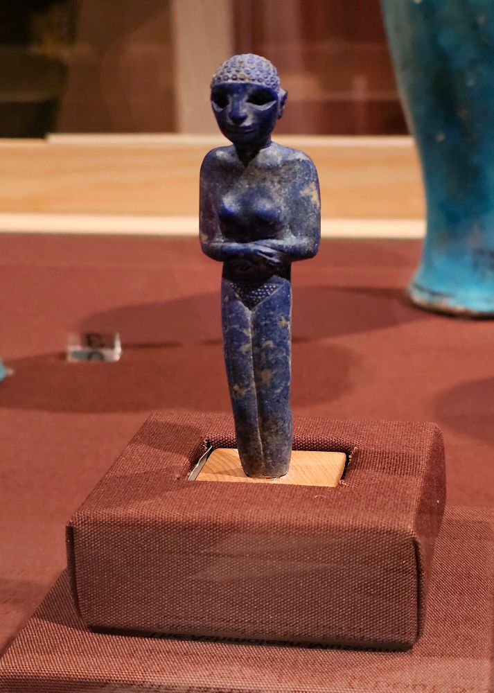 A tiny blue stone figurine with arms crossed standing on a red museum plinth