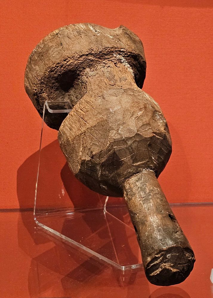 A hefty carved and dented wooden mallet resting on a museum stand, on a red background